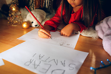 Image showing Merry Christmas and Happy Holidays. Cute little child girl writes the letter to Santa Claus near Christmas tree