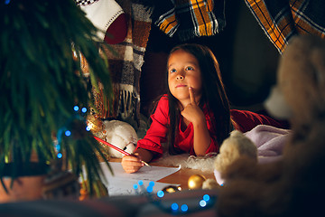 Image showing Merry Christmas and Happy Holidays. Cute little child girl writes the letter to Santa Claus near Christmas tree