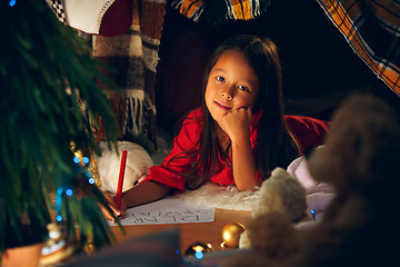 Image showing Merry Christmas and Happy Holidays. Cute little child girl writes the letter to Santa Claus near Christmas tree