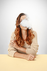 Image showing Tranquil woman sitting and smoking resting at the table. Cloud of smoke covering her face. Copy space