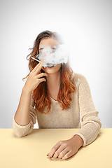 Image showing Tranquil woman sitting and smoking resting at the table. Cloud of smoke covering her face. Copy space