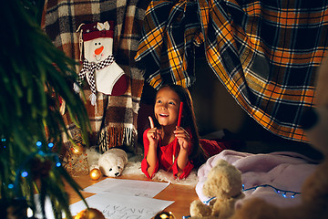 Image showing Merry Christmas and Happy Holidays. Cute little child girl writes the letter to Santa Claus near Christmas tree