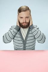 Image showing portrait of stressed man sitting at table with closed and covering her ears with hands.