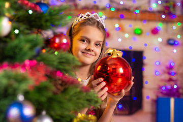 Image showing Happy girl peeps out from behind a beautiful Christmas tree with a ball in her hands