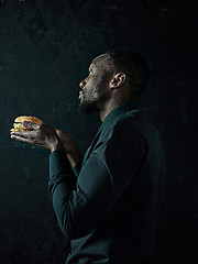 Image showing young african american man eating hamburger and looking away on black studio