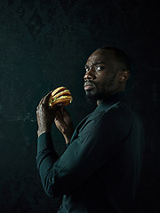 Image showing young african american man eating hamburger and looking away on black studio