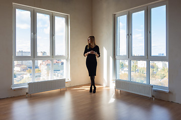 Image showing A girl in a black dress stands between two large windows in a spacious empty apartment