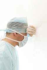 Image showing Tired surgeon in sterile gown and mask leaned his head against the wall