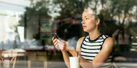 Image showing Thoughtful caucasian woman holding mobile phone while looking through the coffee shop window during coffee break.