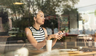 Image showing Thoughtful caucasian woman holding mobile phone while looking through the coffee shop window during coffee break.