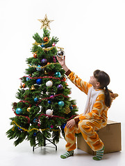 Image showing The girl sits on a box in front of the Christmas tree and hangs a Christmas tree ball