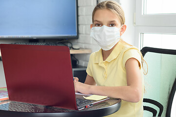 Image showing A girl in a medical mask in a home setting learns remotely and looked into the frame