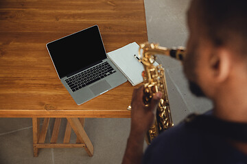 Image showing African-american musician playing saxophone during online concert at home isolated and quarantined. Blank laptop screen with copyspace.