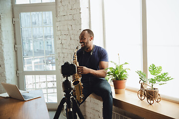 Image showing African-american musician playing saxophone during online concert at home isolated and quarantined, laughting