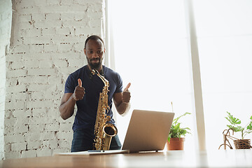 Image showing African-american musician playing saxophone during online concert at home isolated and quarantined