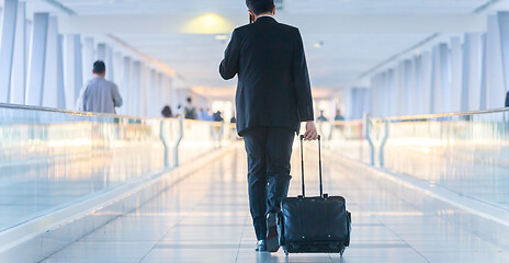 Image showing Businessman walking and wheeling a trolley suitcase at the lobby, talking on a mobile phone. Business travel concept.