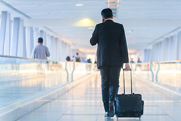 Image showing Businessman walking and wheeling a trolley suitcase at the lobby, talking on a mobile phone. Business travel concept.