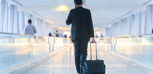 Image showing Businessman walking and wheeling a trolley suitcase at the lobby, talking on a mobile phone. Business travel concept.