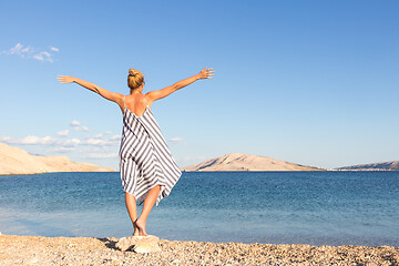 Image showing Happy carefree woman rising arms, wearing beautiful striped summer dress enjoying late afternoon on white pabbled beach on Pag island, Croatia