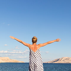 Image showing Happy carefree woman rising arms, wearing beautiful striped summer dress enjoying late afternoon on white pabbled beach on Pag island, Croatia