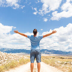 Image showing Rear view of casual sporty man standing on a dirt country road rising hands up to the clouds on a blue summer sky. Freedom and travel adventure concept.