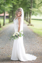 Image showing Full length portrait of beautiful sensual young blond bride in long white wedding dress and veil, holding bouquet outdoors in natural background