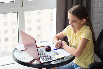 Image showing Girl joyfully looks at the laptop screen while sitting at a table in the apartment and doing homework