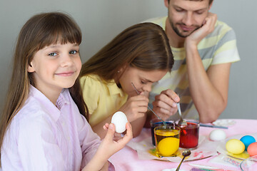 Image showing A girl with an egg in her hands looked into the frame, in the background the family paints eggs in glasses with dyes