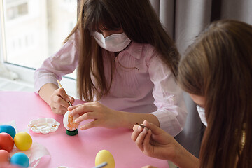 Image showing Two girls in medical masks paint eggs with a brush while preparing for Easter