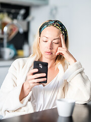 Image showing Beautiful caucasian woman at home, feeling comfortable wearing white bathrobe, taking some time to herself, drinking morning coffee and reading news on mobile phone device in the morning