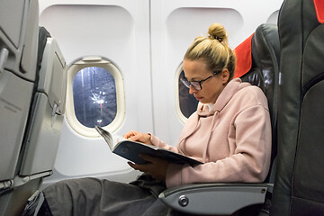 Image showing Casual young woman reading magazine and listening to music on airplane. Female traveler reading seated in passanger cabin.