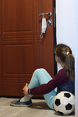 Image showing A girl sits in the hallway and looks at the door, on the handle of which a protective medical mask is hanging