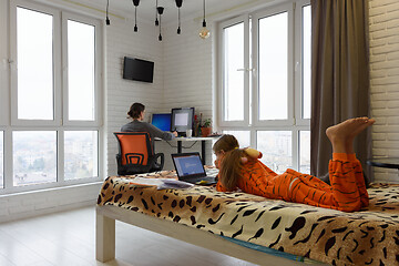 Image showing The family is at home, the child lies on the bed and studies in a laptop, the mother in the background works remotely in the computer