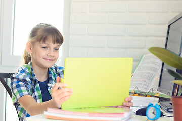 Image showing A girl sits at a table and happily looks at the screen of a tablet computer