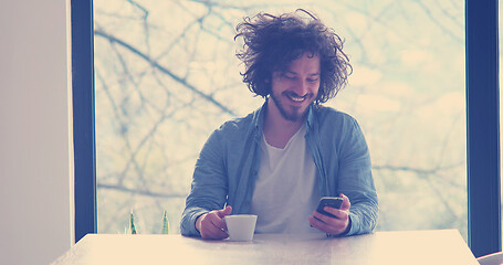 Image showing young man drinking coffee and using a mobile phone  at home