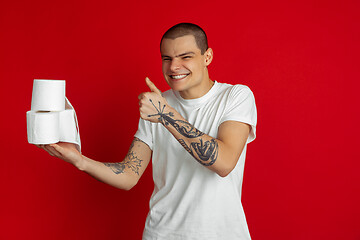 Image showing Caucasian young man\'s portrait on red studio background - holding toilet papers, essential goods during quarantine and self-insulation