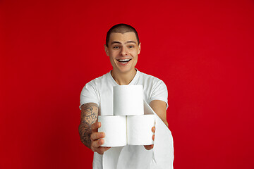 Image showing Caucasian young man\'s portrait on red studio background - holding toilet papers, essential goods during quarantine and self-insulation