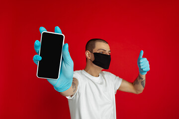 Image showing Caucasian young man in protective face mask and medical gloves on red studio background - showing blank phone\'s screen