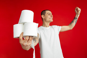 Image showing Caucasian young man\'s portrait on red studio background - holding toilet papers, essential goods during quarantine and self-insulation