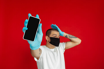 Image showing Caucasian young man in protective face mask and medical gloves on red studio background - showing blank phone\'s screen