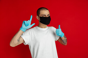 Image showing Caucasian young man in protective face mask and medical gloves on red studio background - pointing and showing thumb up