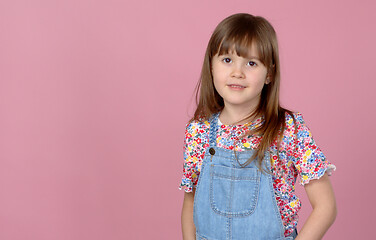 Image showing Sweet little girl 6-7 years old posing in dungarees jeans and flower pattern blouse on pink