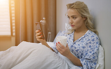 Image showing Beautiful young woman browsing smartphone and holding mug of hot beverage sitting on bed