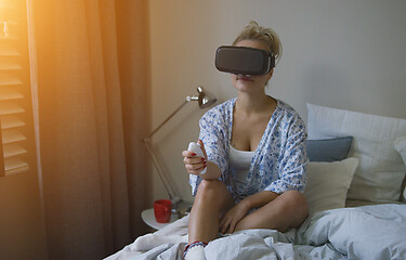 Image showing Young woman in shirt and VR headset holding controller while sitting on bed