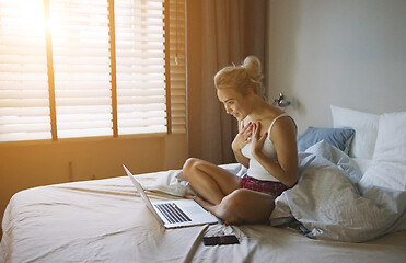 Image showing Charming woman in pajamas sitting on bed near laptop and looking at it