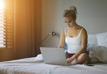 Image showing Pretty young woman in shorts and tank top sitting on comfortable bed and using laptop