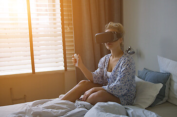 Image showing Young woman in shirt sitting on comfortable bed and using VR headset with controller