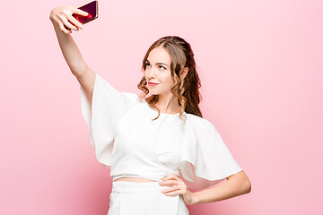 Image showing Portrait of a young attractive woman making selfie photo with smartphone on a pink background