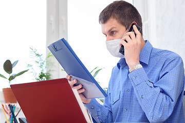 Image showing A man with a medical mask on his face reads a document in the office and talks on the phone