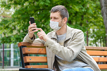 Image showing A man in a medical mask with a smartphone sits on a bench in the park